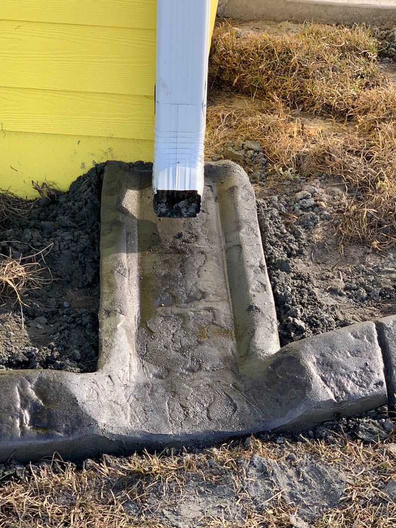 A white gutter downspout emptying into a concrete splash block on yellow siding, outdoors.
