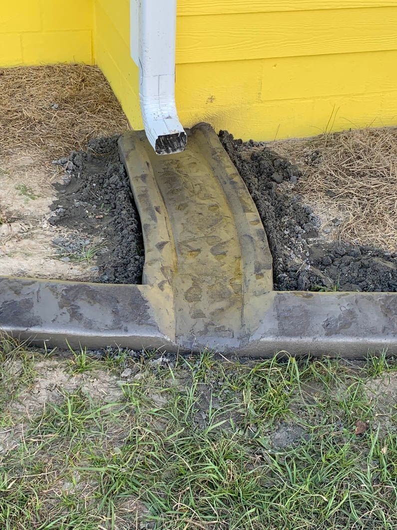 Yellow building corner with a downspout draining onto a concrete splash block. Grass in the foreground.