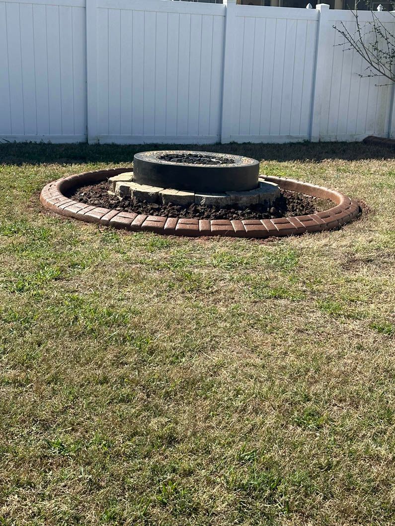 Fire pit with a brick base in a grassy yard, surrounded by a brown, decorative border, white fence in background.