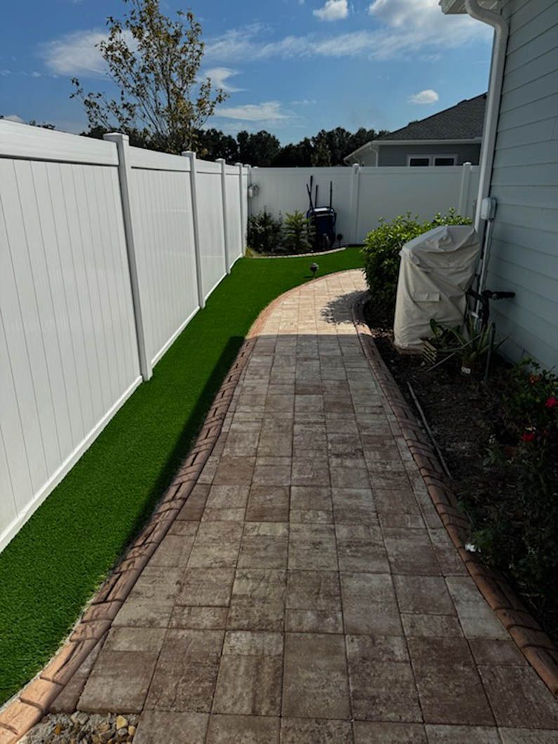 Brick pathway curves along green turf and white fence, beside a house exterior on a sunny day.