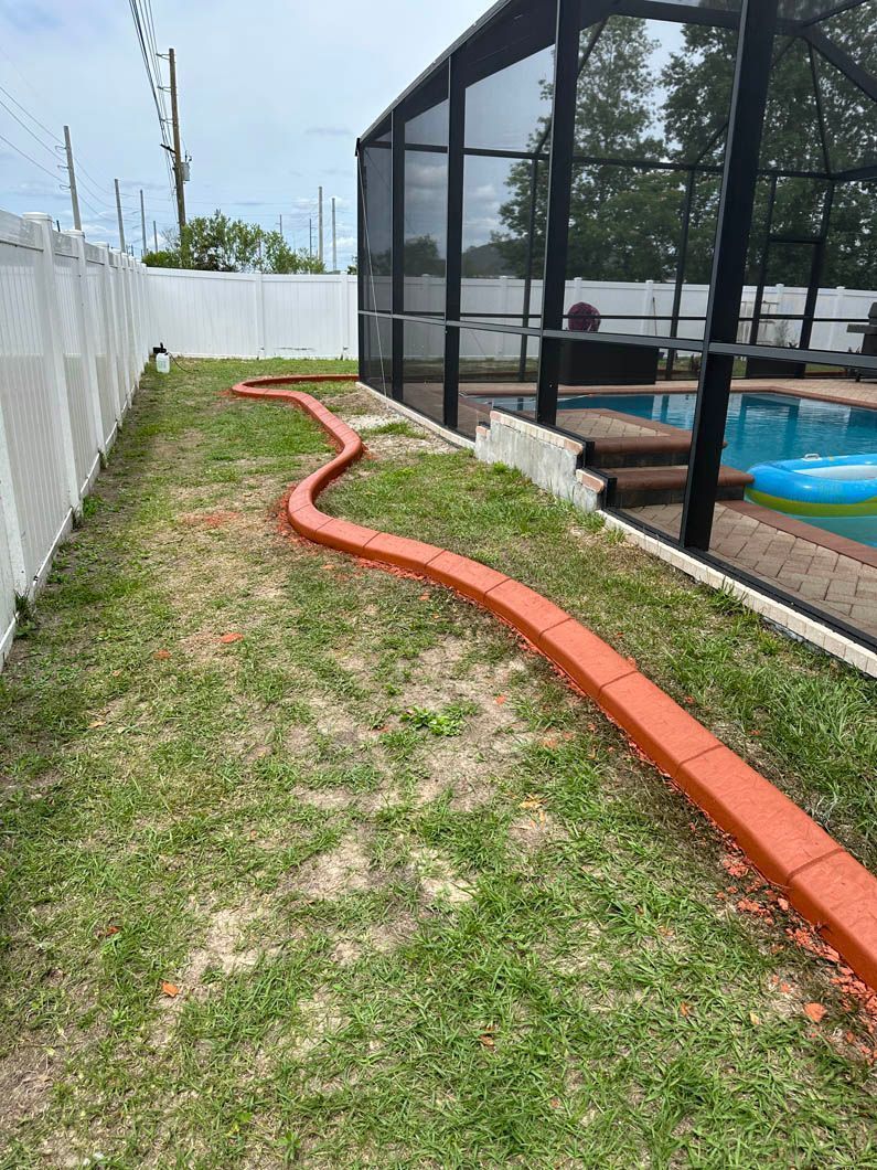 Grassy backyard path lined with orange edging, leading to a swimming pool. White fence on the left, pool enclosure on the right.