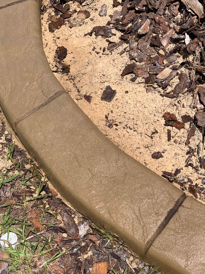 Brown curved concrete curbing bordering a sandy area with mulch.