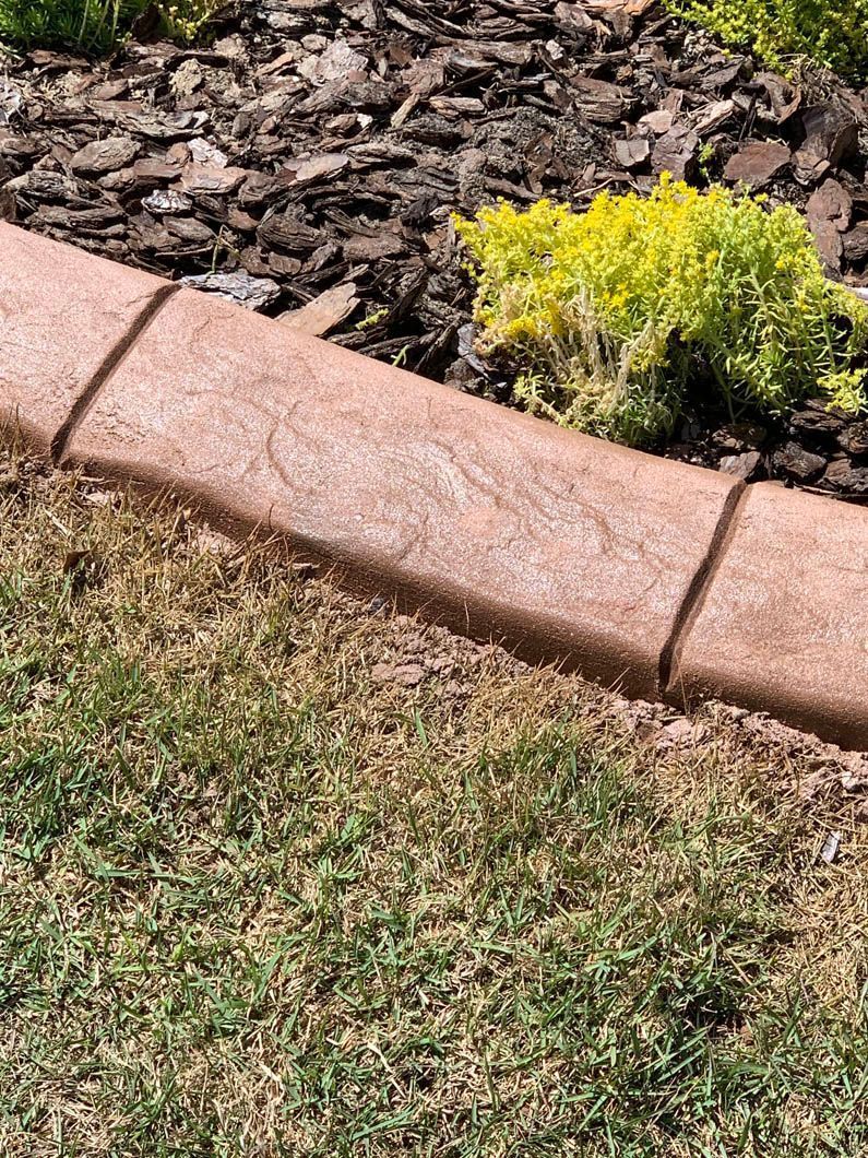Brown decorative concrete curbing borders a garden bed with mulch and green plants.