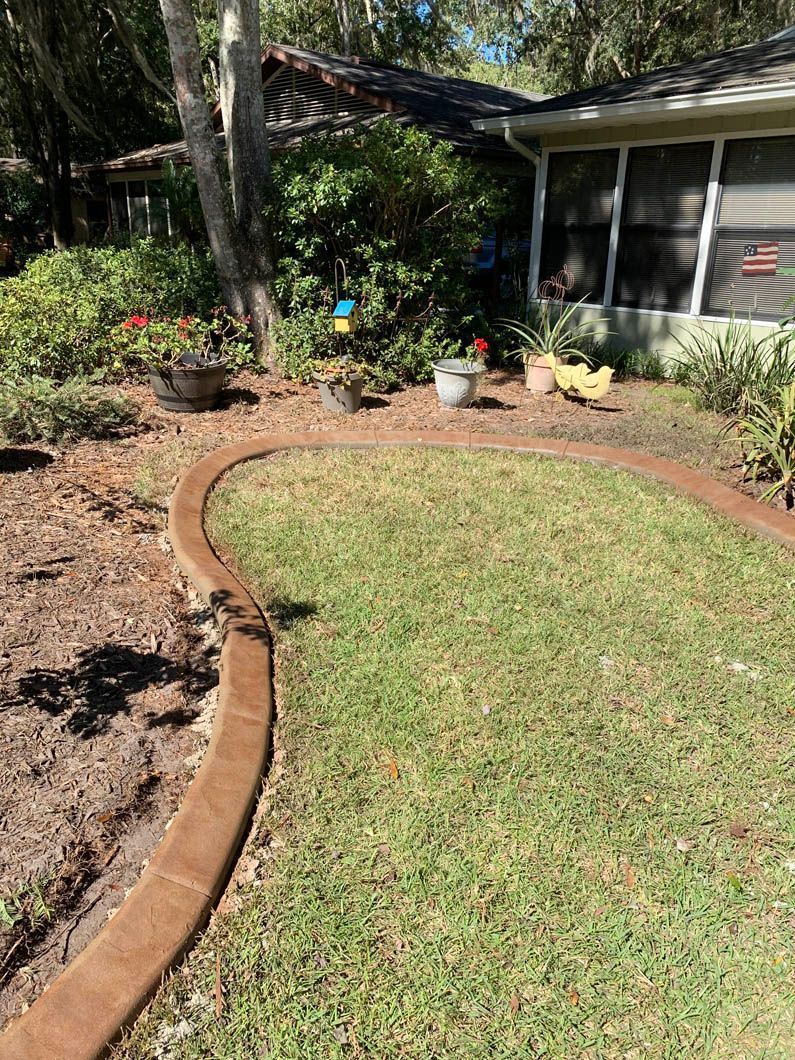 Brown-edged flower bed with green grass, plants, and a house in the background on a sunny day.