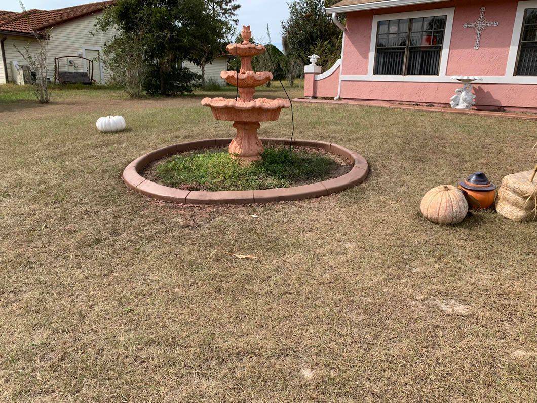 A brown fountain in a lawn with pumpkins, near a pink house.