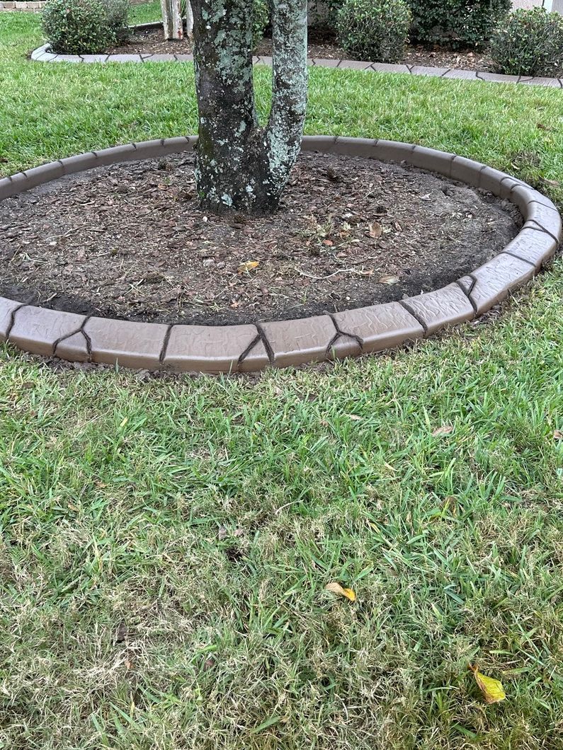 Tree trunk surrounded by brown, stone-like concrete edging in a grassy lawn.