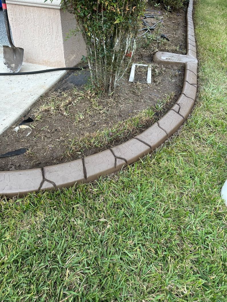 Curved brown concrete curbing around a garden bed with grass and a building.