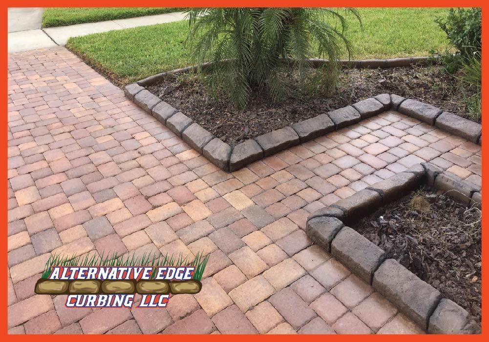 Brick pavers pathway with dark edging, grass, and a plant bed in front of a house.