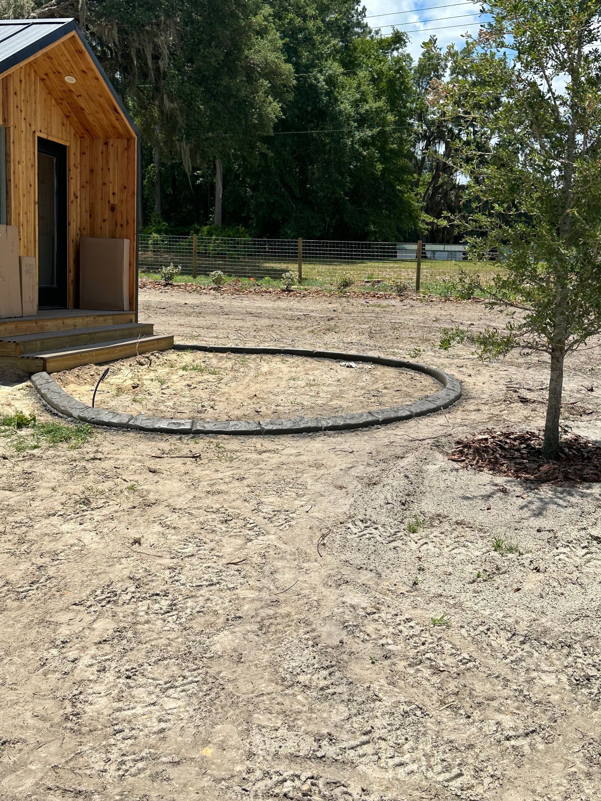 Small wood cabin, curved border of black material, sandy ground. Tree on right. Sunny, outdoor setting.