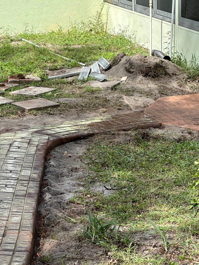 Brick pathway under construction next to dirt pile and scattered paving stones, by a building.