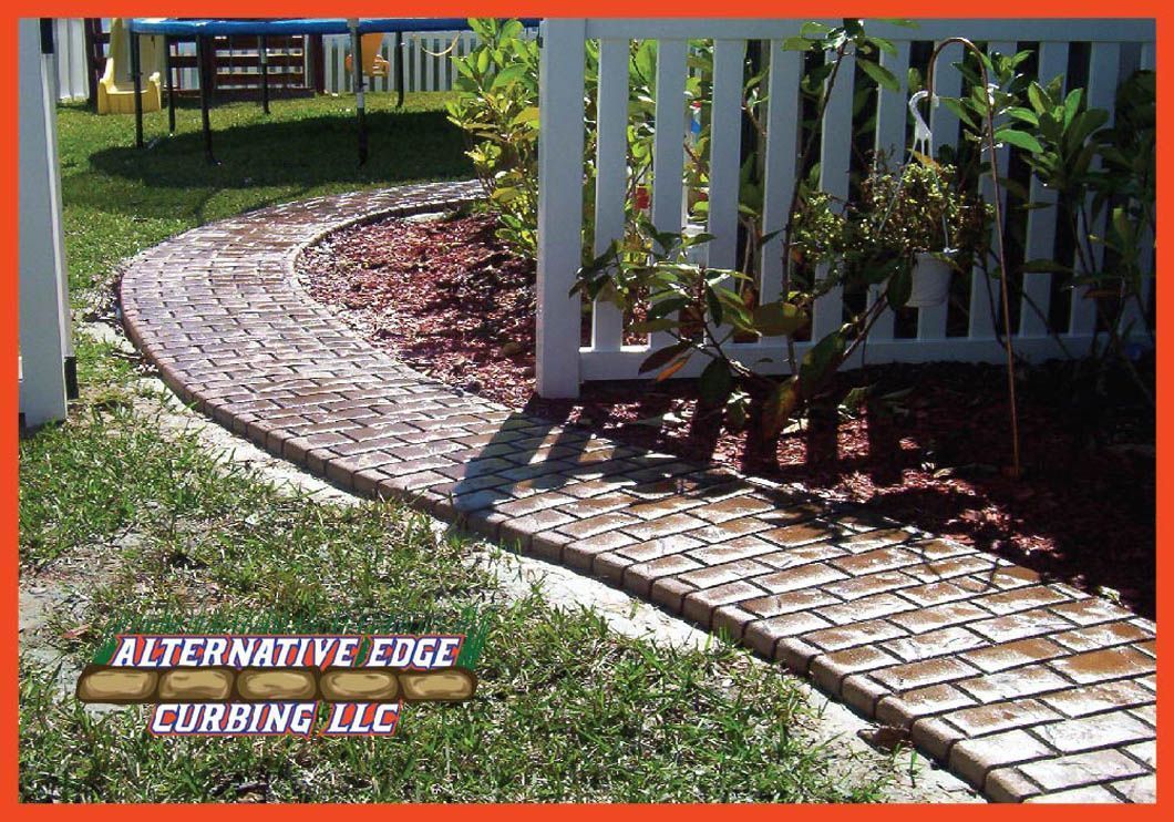 Brick path with curved border alongside a white picket fence and garden.
