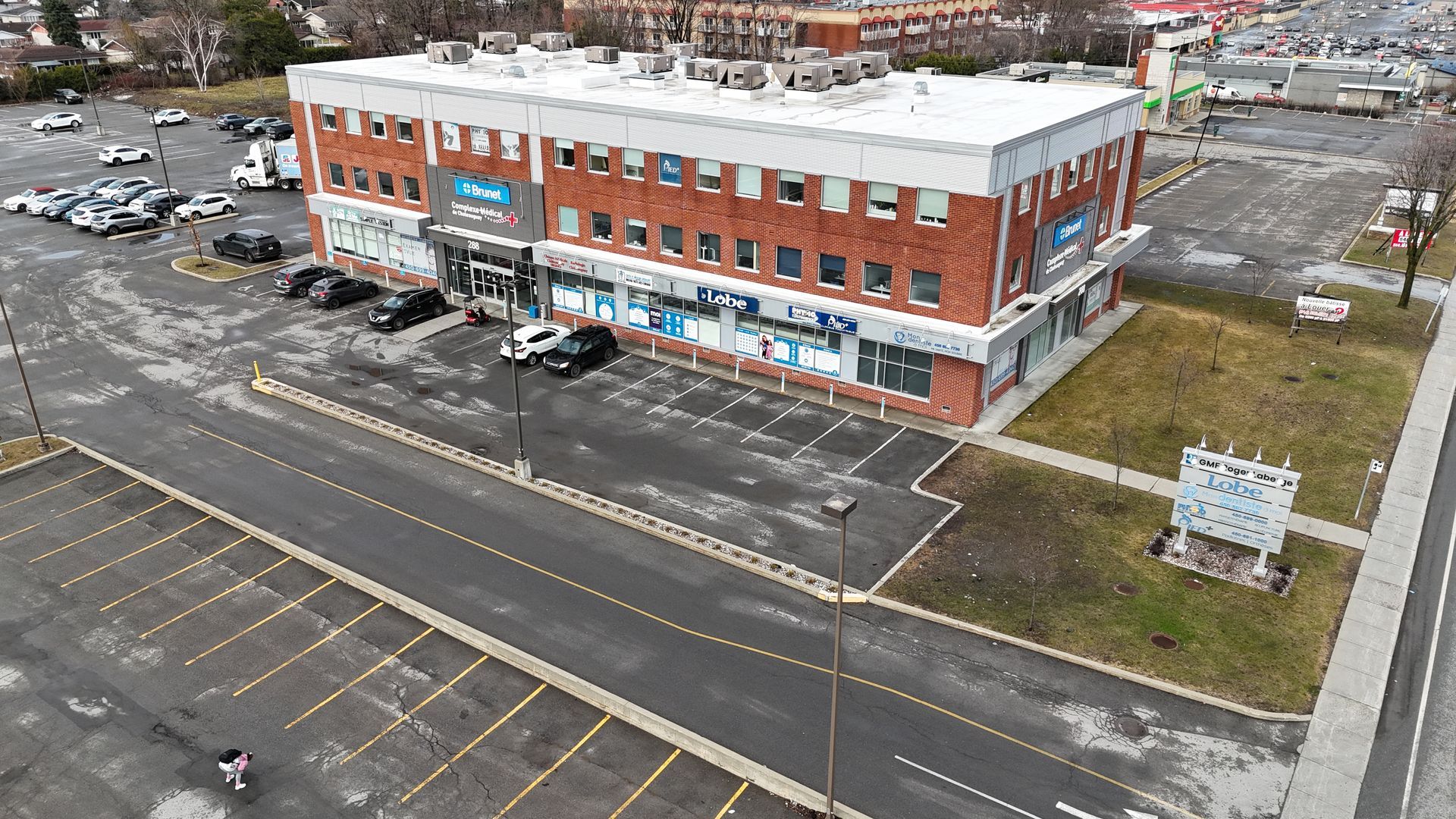 Vue aérienne d'un bâtiment commercial en briques de deux étages avec un parking et les rues environnantes.