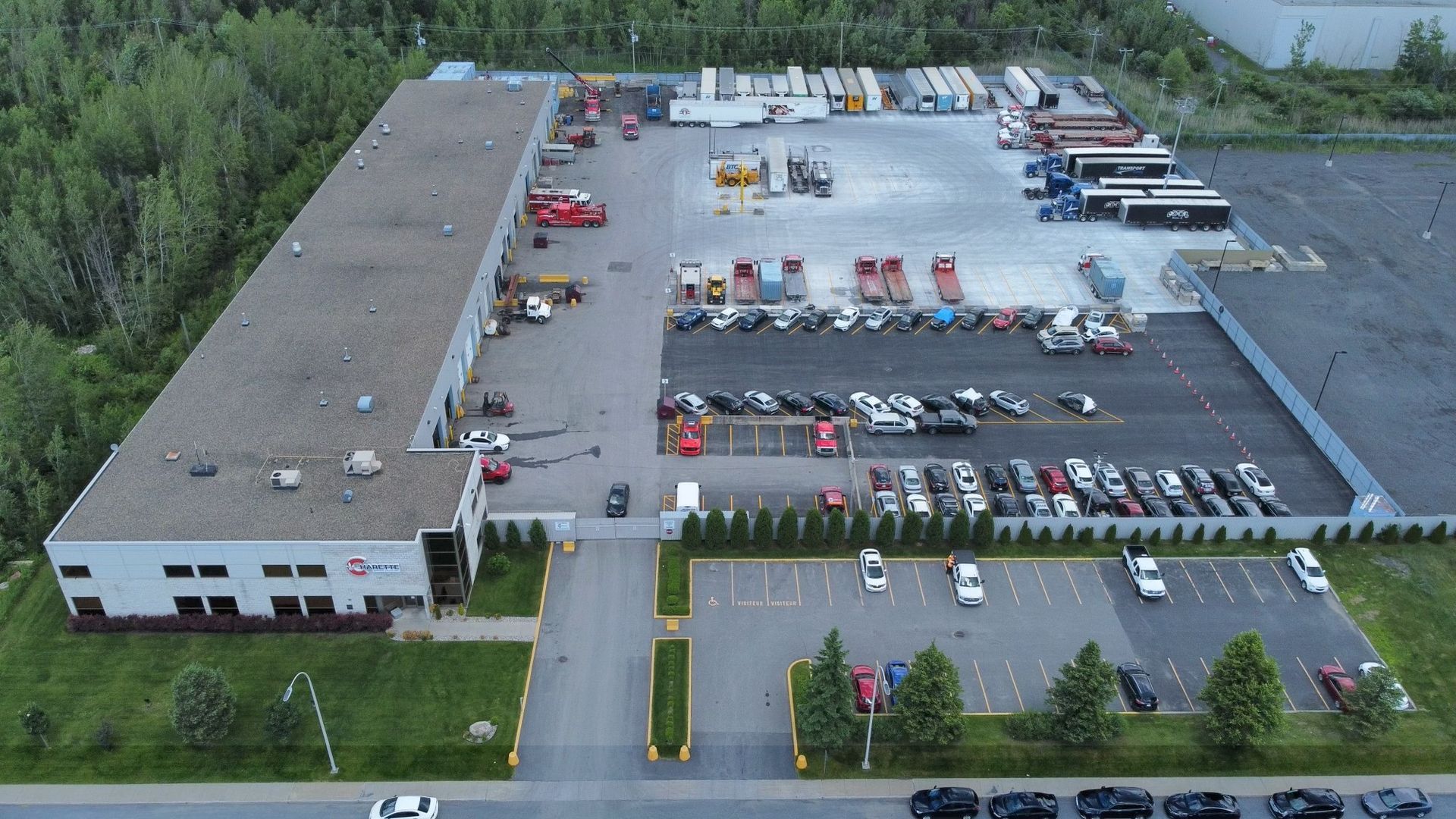 Vue aérienne d'un grand bâtiment industriel avec un parking rempli de camions et de voitures, entouré d'arbres.