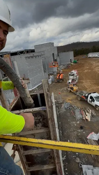 Construction worker pours concrete into a formwork. Construction site with machinery, cloudy sky.