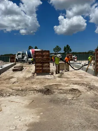 Construction site with workers pouring concrete into forms; blue sky, white clouds.