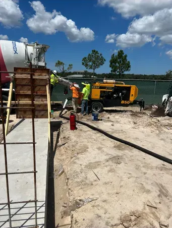 Construction workers pouring concrete at a building site; includes a cement truck, pump, and rebar framework.