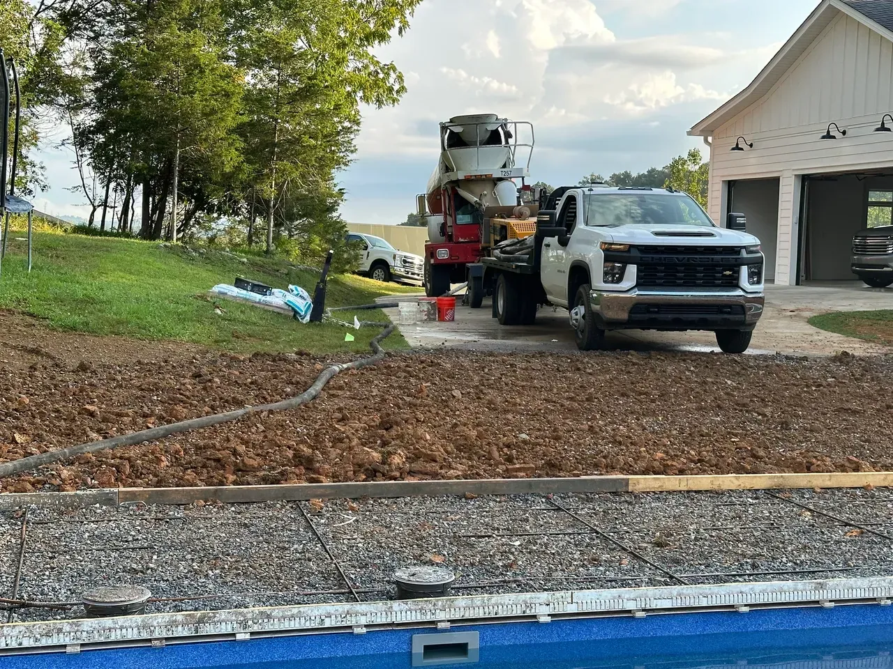 Cement truck pouring concrete by a pool with a truck parked in front of a garage.