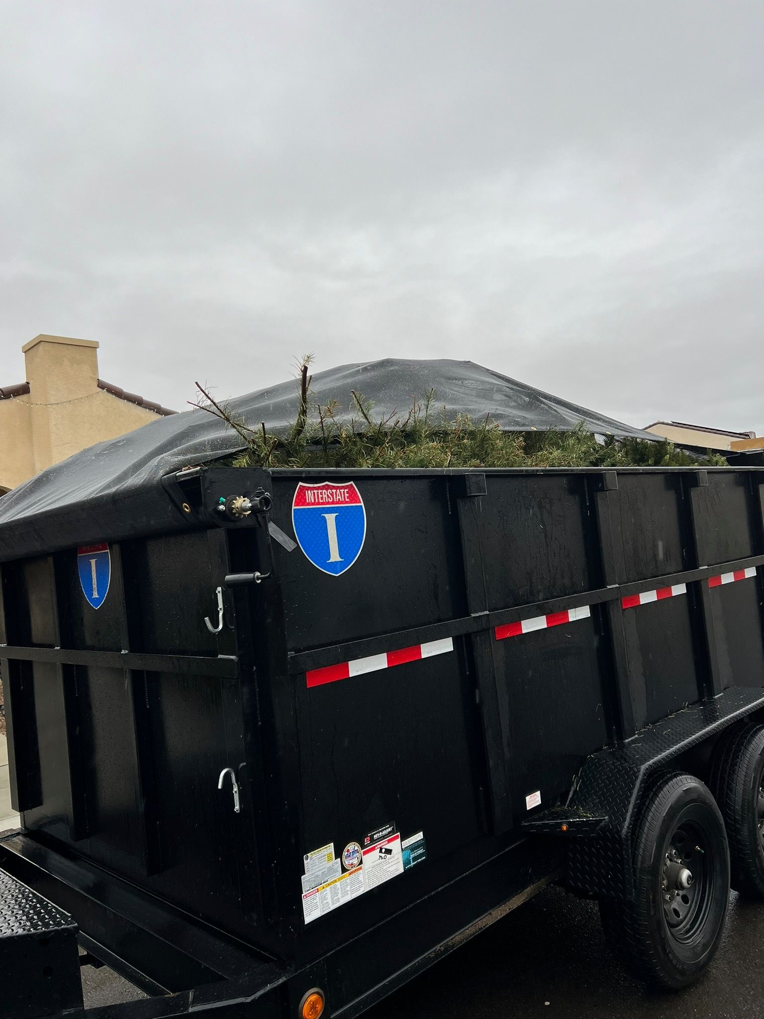 Black trailer filled with green branches, labeled with an Interstate 1 sign on a cloudy day.