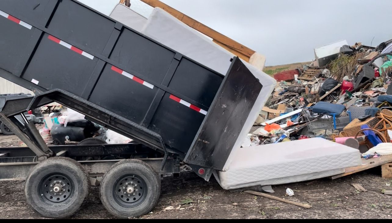 A black dump trailer dumping debris, including a mattress, onto a large pile of trash.