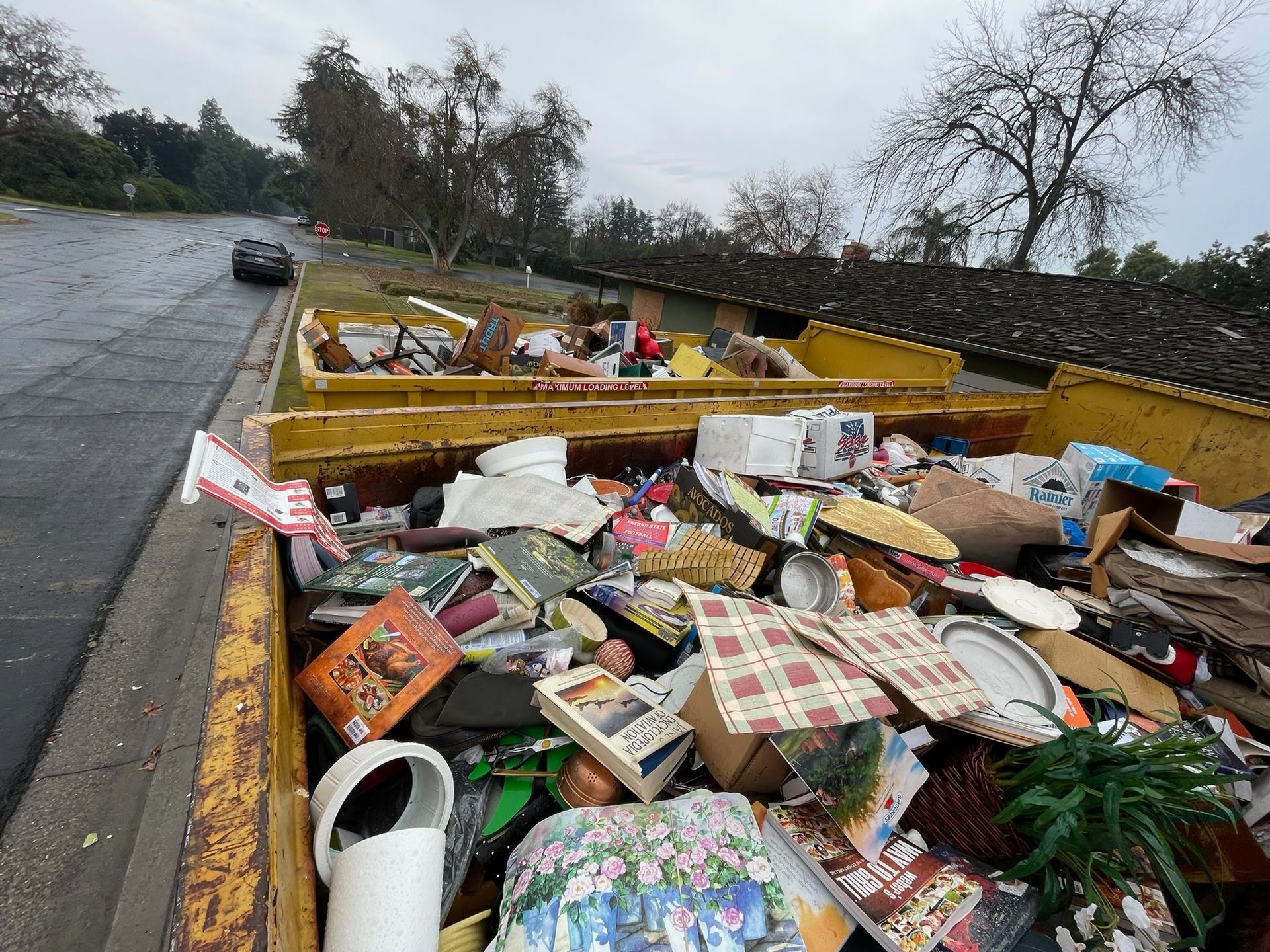 Yellow dumpster overflowing with household trash and debris; outdoors on a cloudy day.
