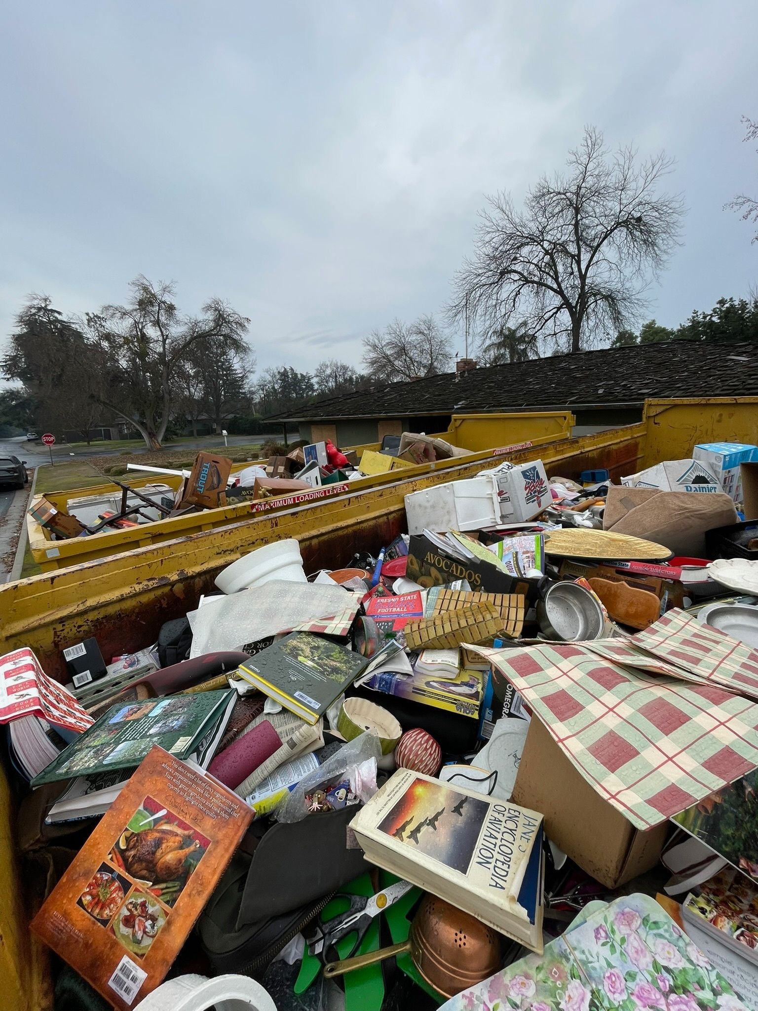 A yellow dumpster overflowing with trash and discarded items outdoors on a cloudy day.