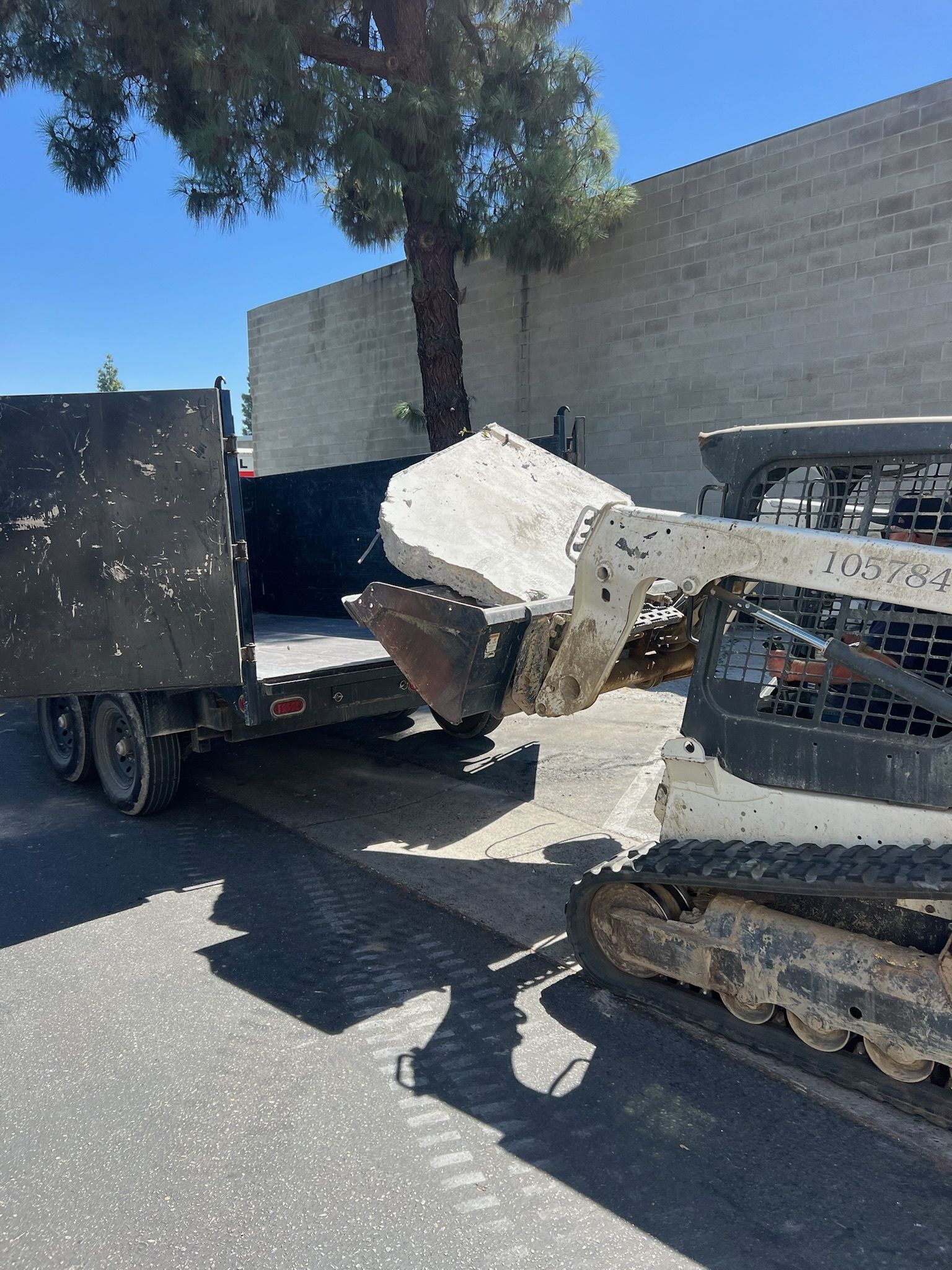 A skid steer loader placing a concrete slab into a trailer on a sunny day.