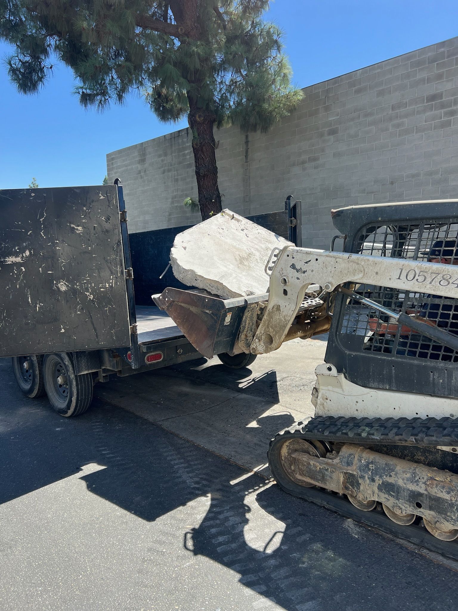 Bobcat loading a large concrete block into a black trailer on a sunny day.