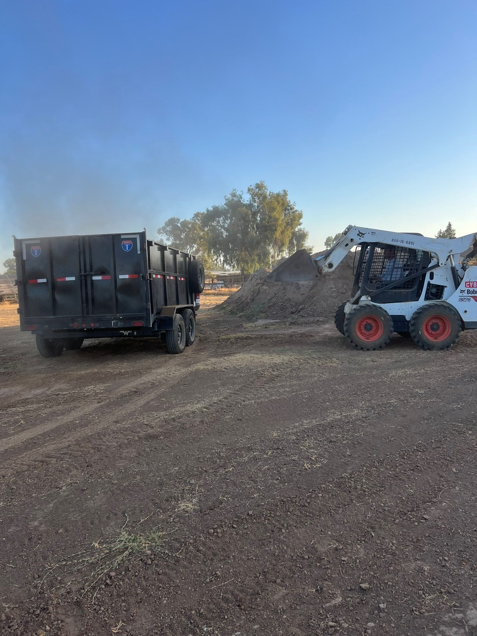 Bobcat loading dirt into a black dump truck on a dusty field under a blue sky.