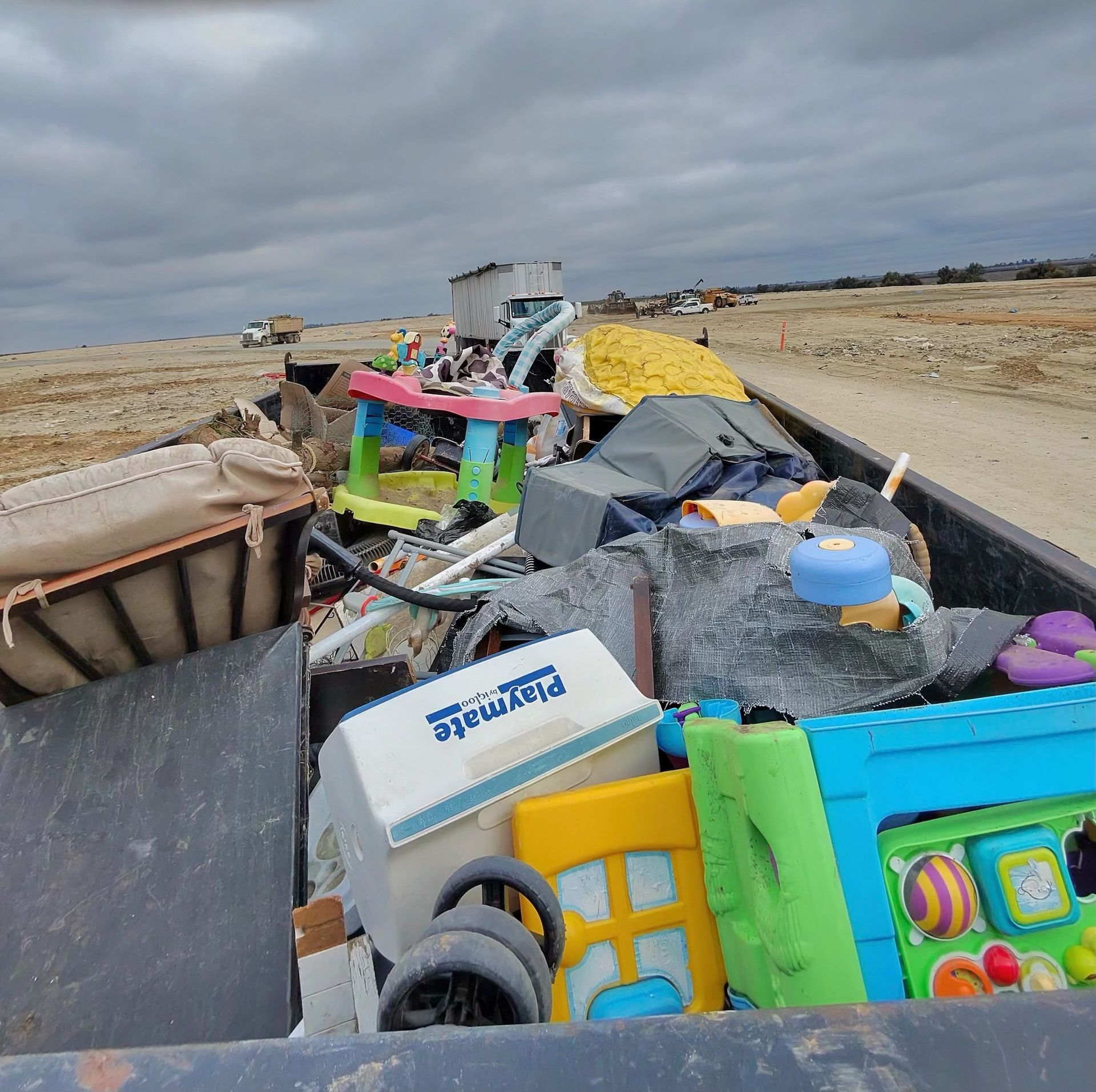 A container filled with trash including children's toys sits outdoors under a cloudy sky.