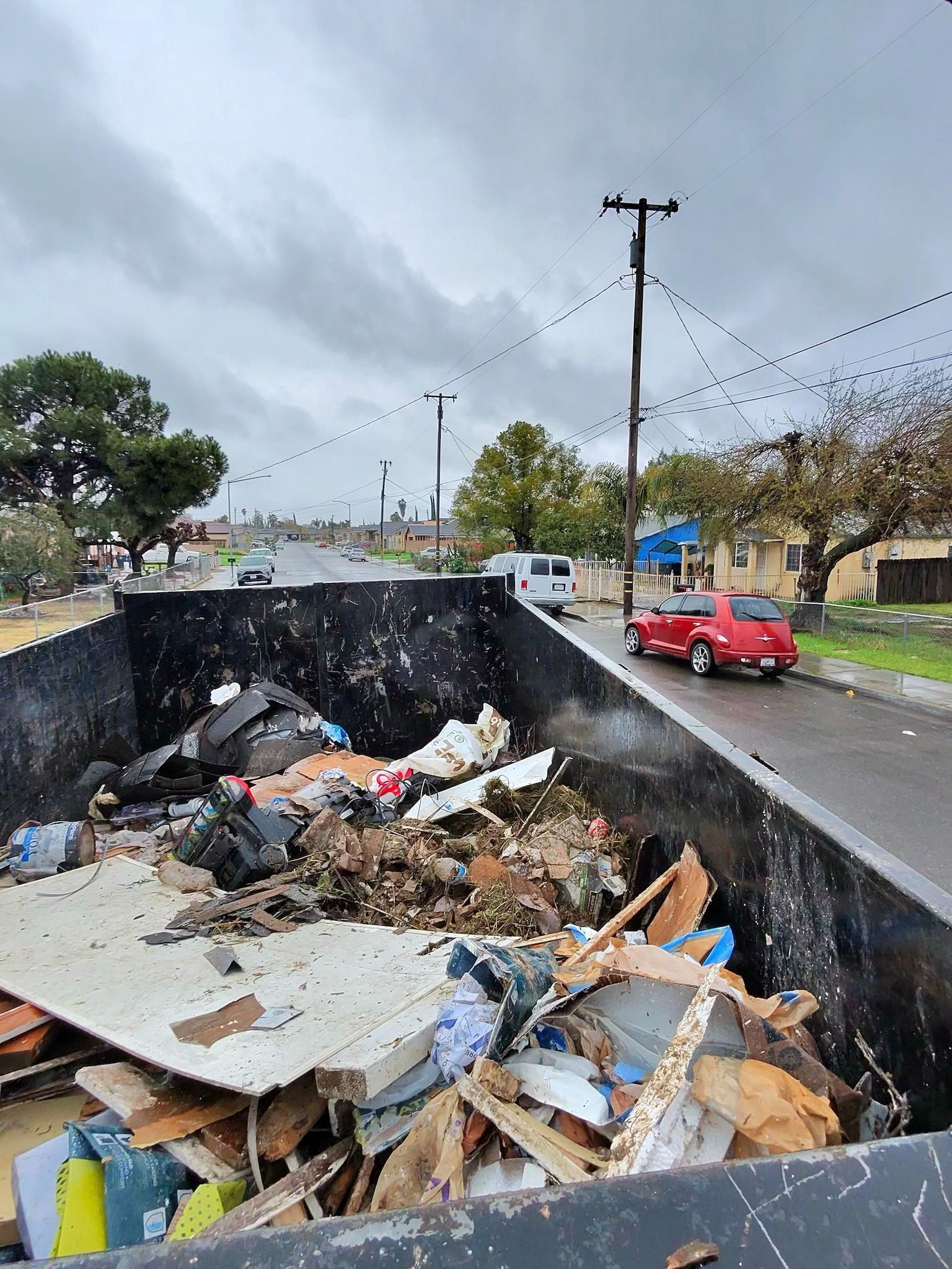 Dumpster overflowing with debris next to a road with a red car driving. Overcast day.