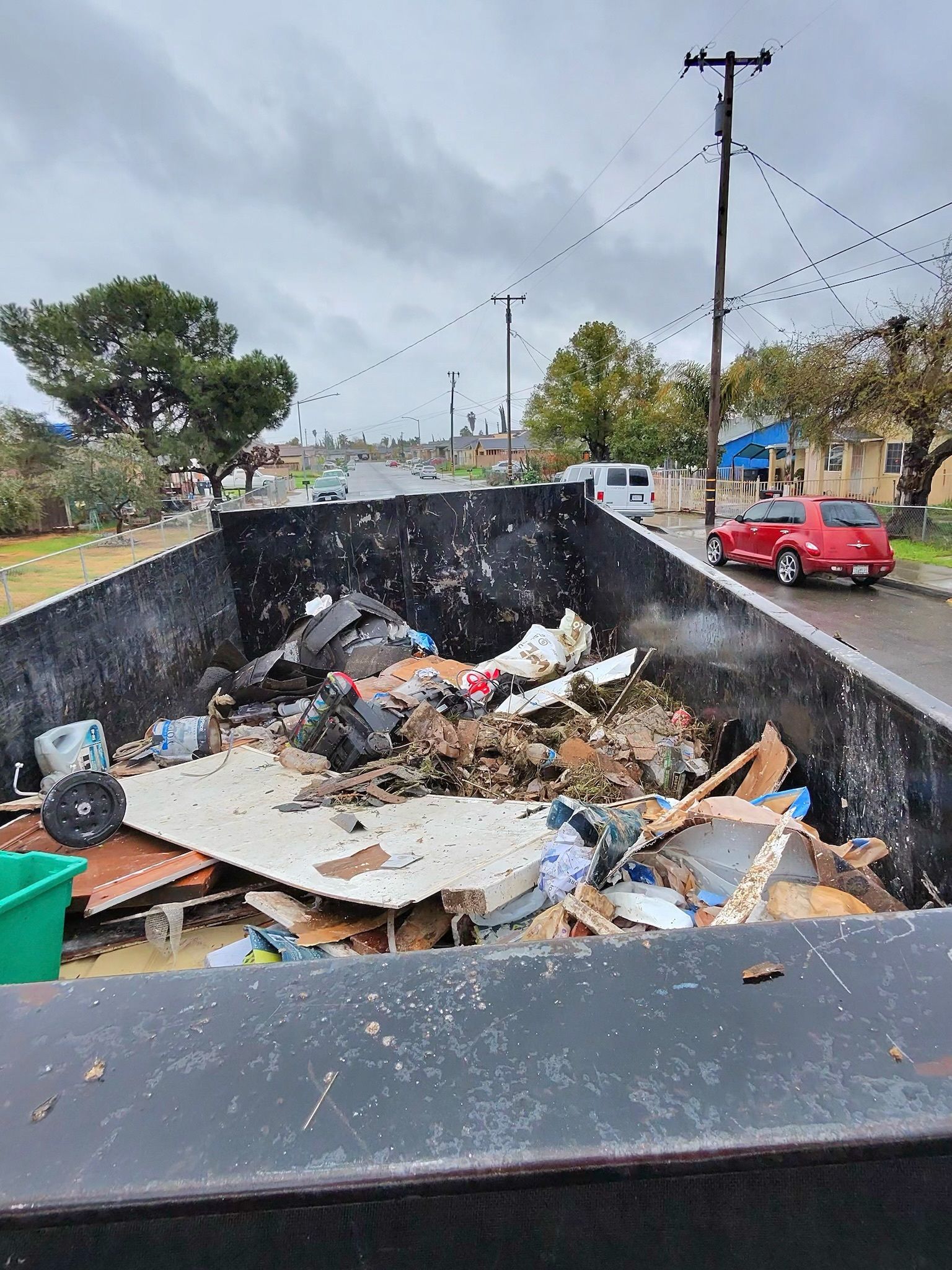 Dumpster filled with debris sits curbside with a red car and overcast sky in the background.