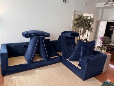 A dark blue modular sofa with some back cushions arranged vertically and horizontally on a light-colored rug in a room.