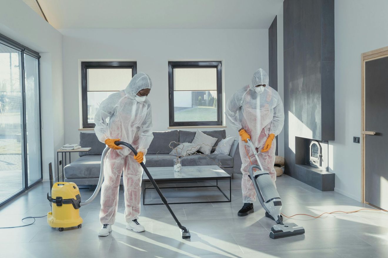 Two people in white protective suits and masks clean the floor of a modern, minimalist living room with vacuum equipment.