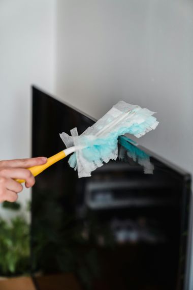 A hand uses a yellow-handled feather duster to clean the top edge of a black television screen against a white wall.