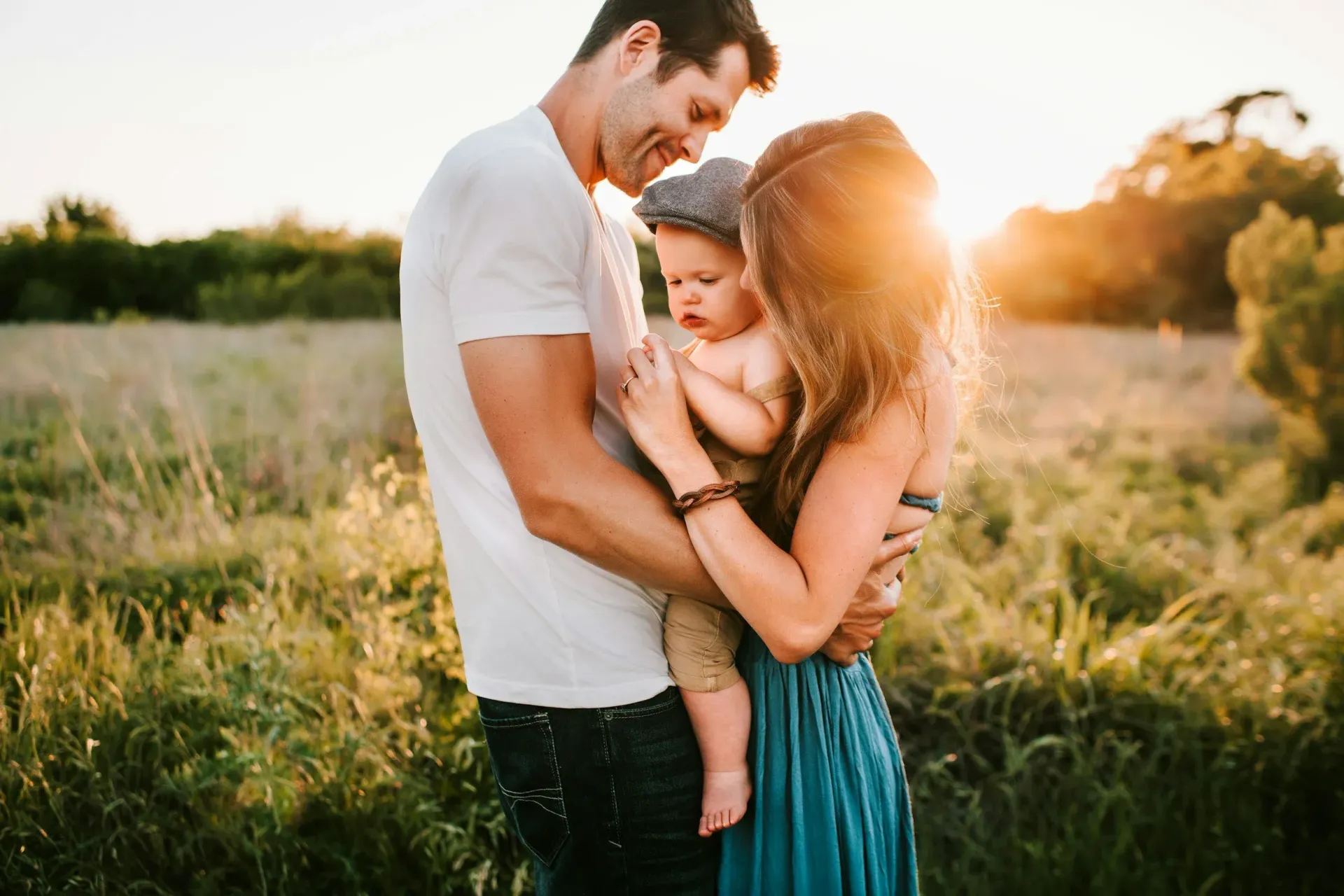 A family of three embrace in a field during a golden sunset, with the parent in the middle wearing a flat cap.