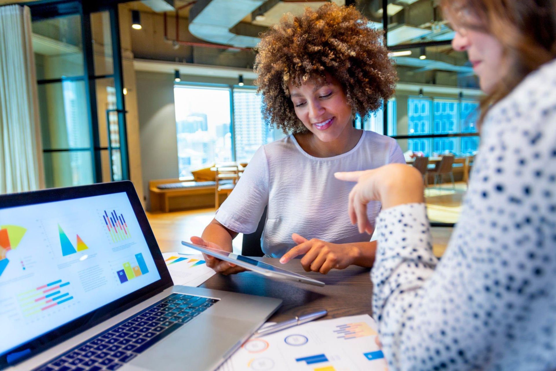 Two women are sitting at a table looking at a laptop and a tablet.