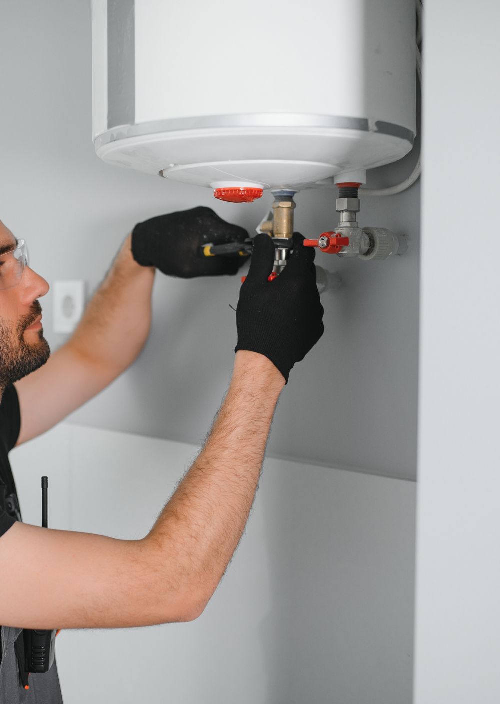 A worker in black gloves repairs a white water heater mounted on a grey wall.