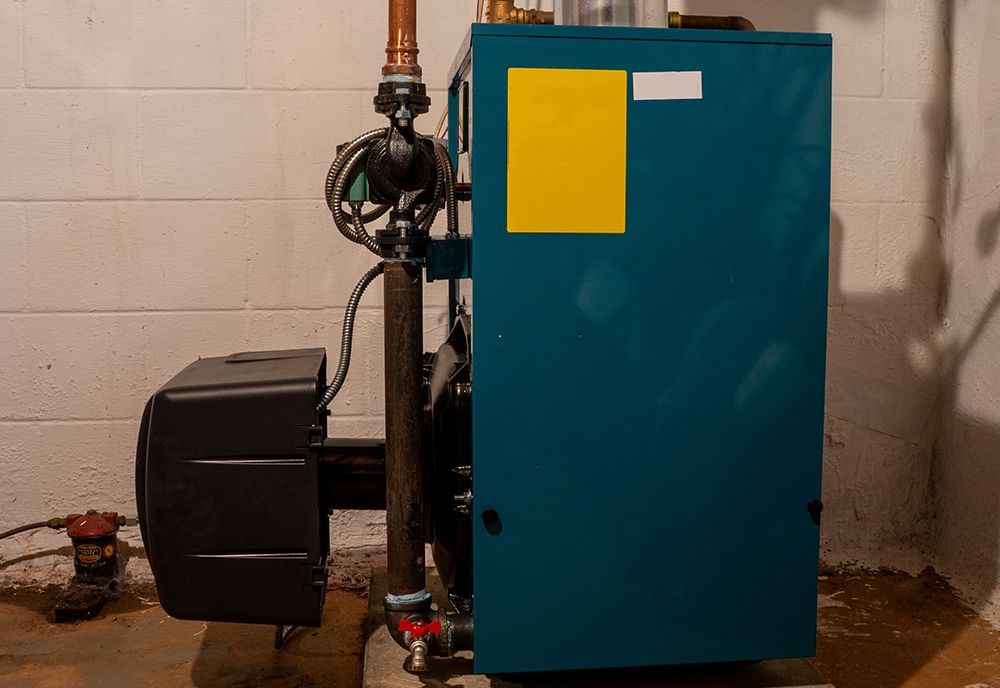 A blue oil-fired residential boiler with attached copper piping and a circulator pump stands against a white block wall.