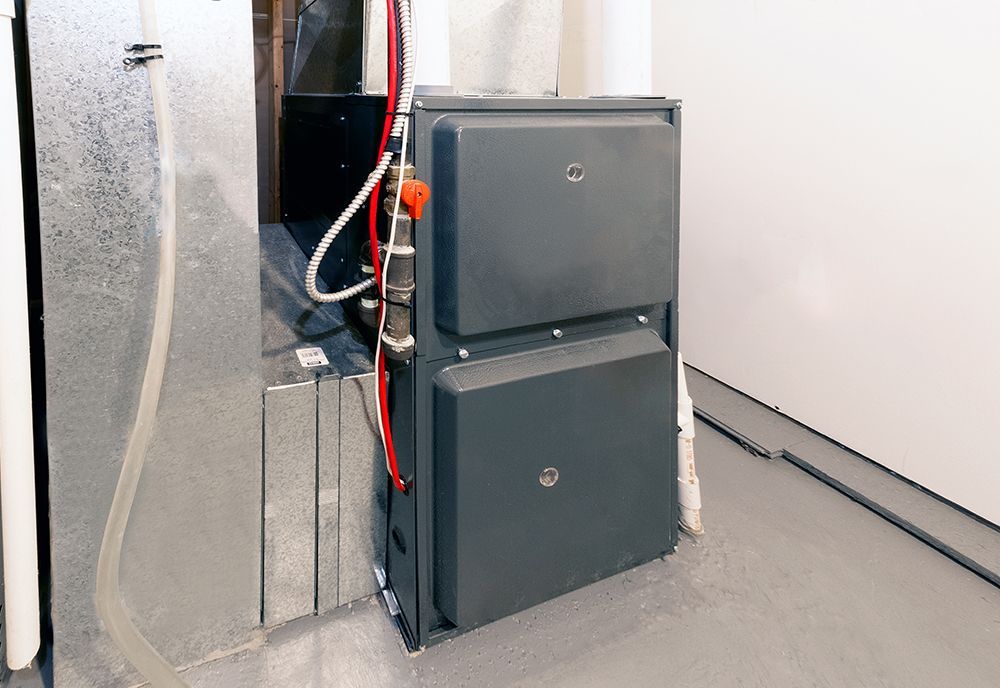 A gray heating furnace unit sits on a concrete floor next to silver metal air ducts.