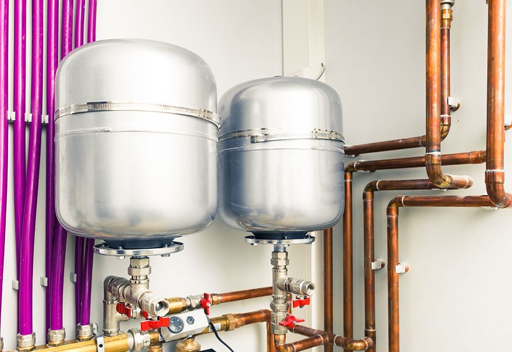 Two metallic expansion tanks connected to a network of copper pipes against a white wall with vertical purple conduits.