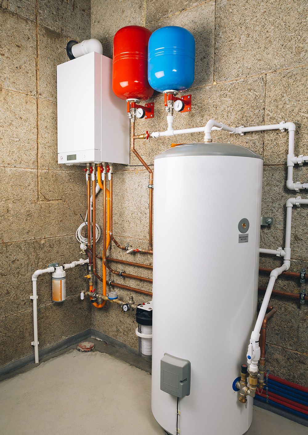 A white boiler and hot water cylinder installed against a textured block wall with red and blue expansion vessels above.