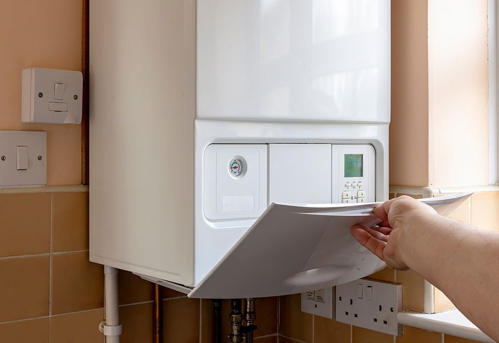 A hand opens the front panel of a white wall-mounted boiler in a kitchen with tan tiled walls.