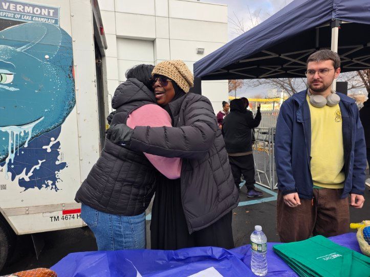 Two women hugging near a table; a man watches with headphones around his neck; an event outdoors.