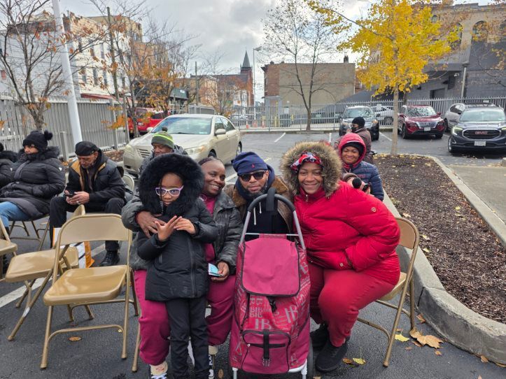 Group of people outdoors near parked cars, some seated in chairs, smiling, bundled in winter coats.