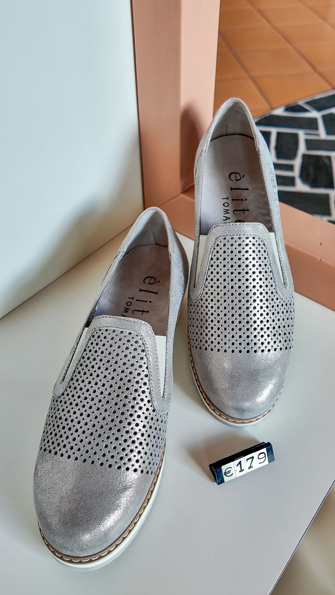 Scarpe slip-on argentate con dettagli traforati, su una superficie bianca. Una piccola etichetta è lì vicino.