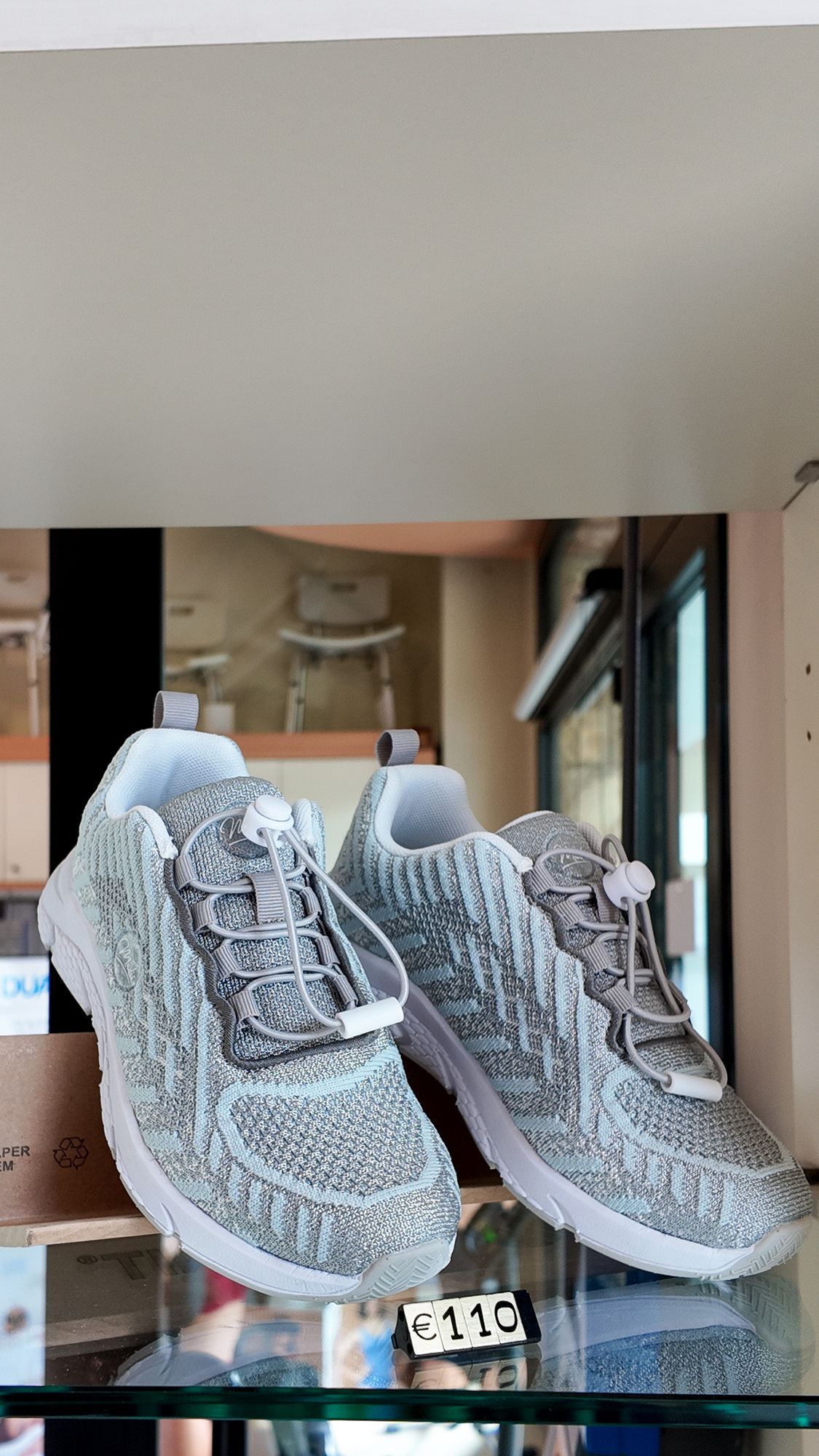 Scarpe sportive bianche e argentate con lacci elastici esposte su uno scaffale di vetro.