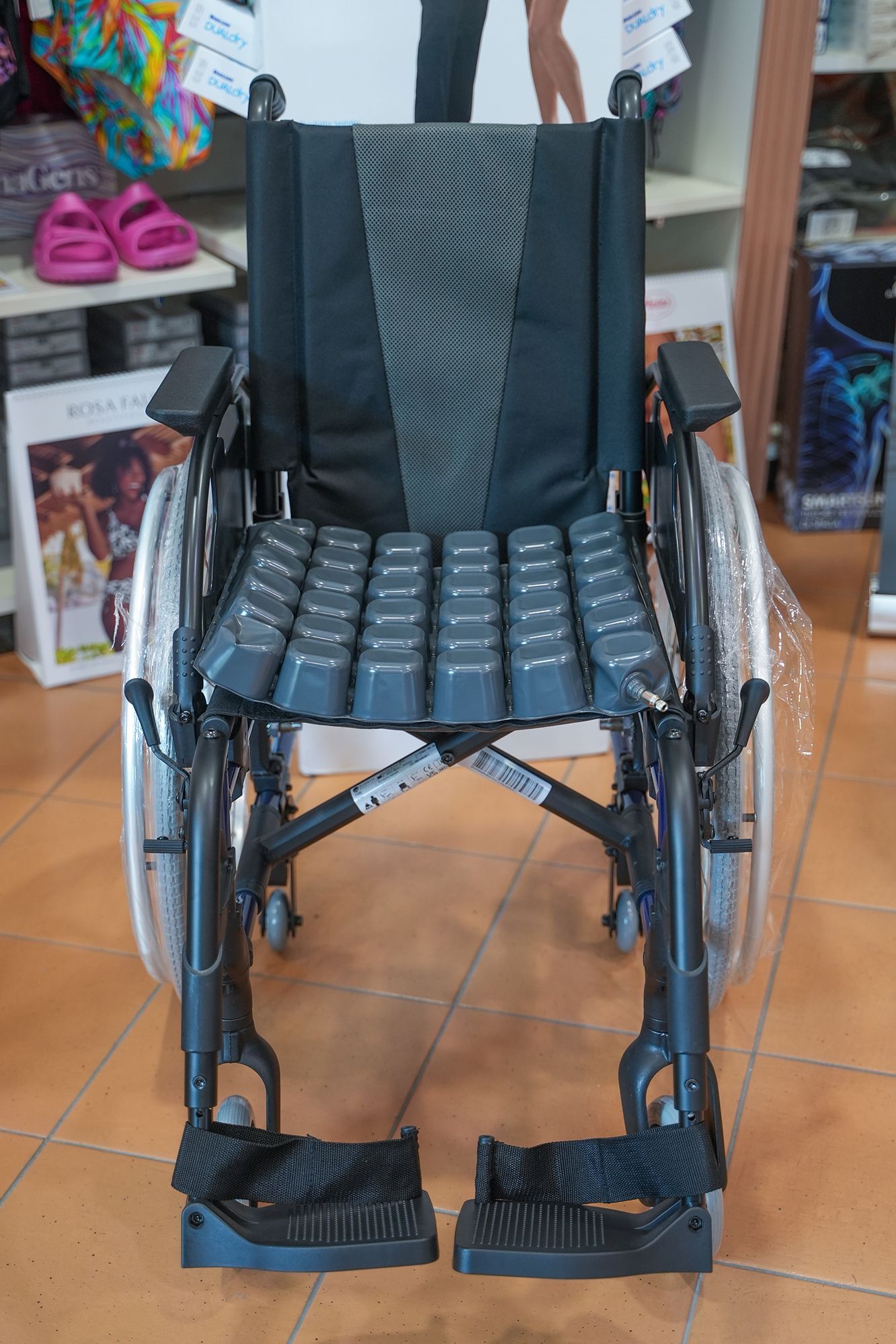 Sedia a rotelle nera con sedile e schienale imbottiti, poggiapiedi e grandi ruote posteriori, posizionata su un pavimento piastrellato.