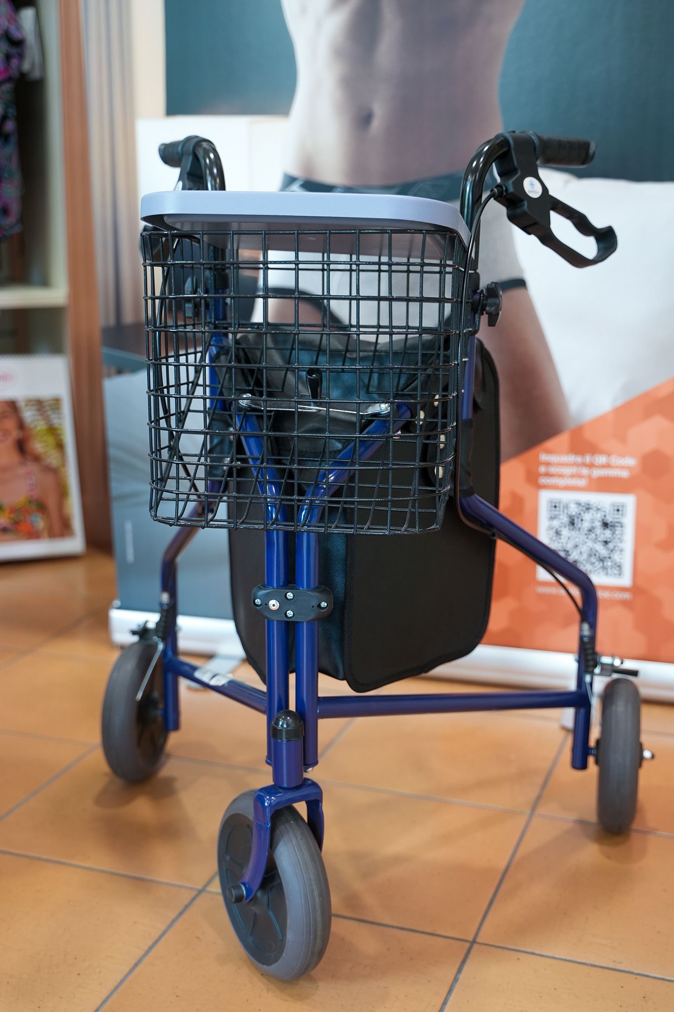 Deambulatore blu a tre ruote con cestino e borsa nera, in un ambiente interno, su un pavimento piastrellato, possibilmente in un negozio.
