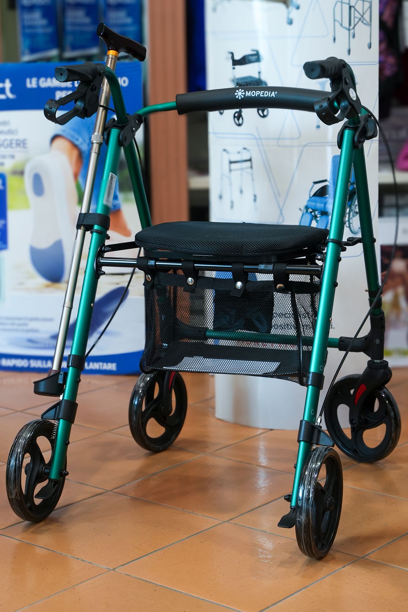 Deambulatore verde con seduta nera e cestino portaoggetti, in casa su pavimento piastrellato.