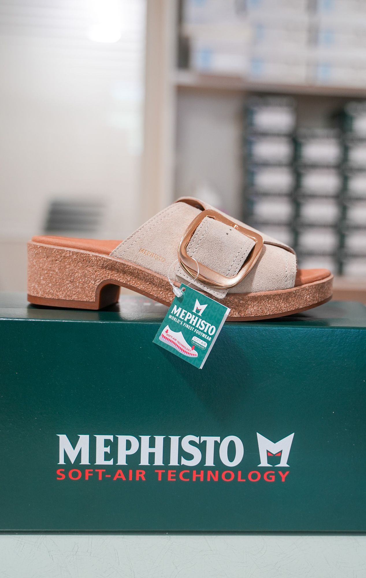 Sandalo Mephisto beige con fibbia grande, sopra una scatola Mephisto verde.